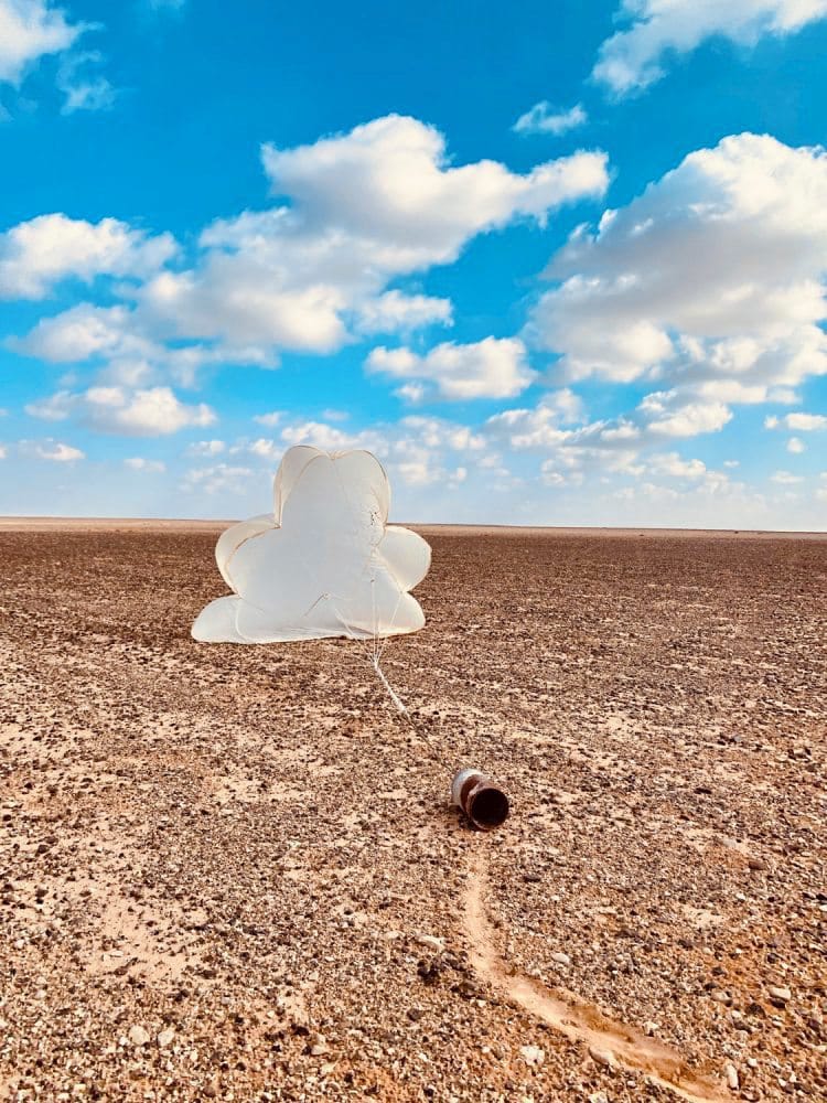 Leere, rostige Konservendose auf einem Feld, an der ein sich wolkenförmig aufblähender Fallschirm befestigt ist, vor einem blauen Himmel mit Wolken