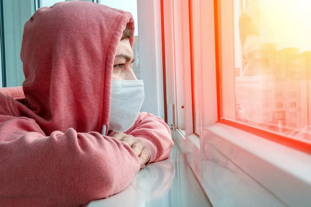 Junger Mann sitzt mit Maske vor dem Fenster