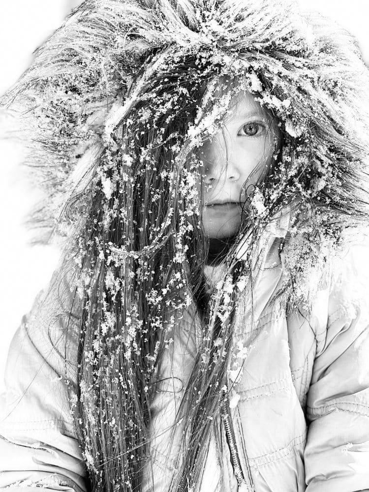 In schwarz-weiß fotografiertes, eingeschneites Mädchen in Felljacke mit hochgezogener Kapuze