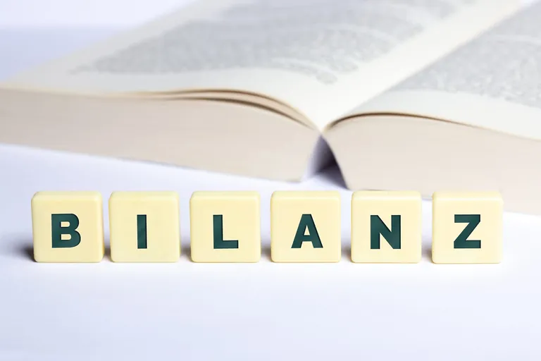 Bilanz in Buchstaben auf W&uuml;rfel mit ge&ouml;ffnetem Buch im Hintergrund.