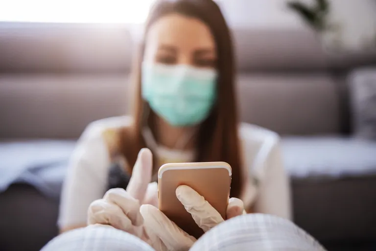 Frau mit Maske in Wohnzimmer tippt auf Handy.