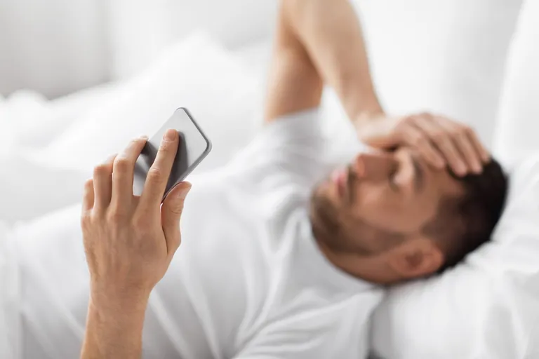 Mann mit Kopfschmerzen im Bett, h&auml;lt Smartphone in der Hand.