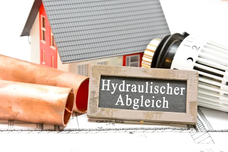 Miniaturhaus, Heizungsrohre, Thermostat und Tafel mit der Aufschrift "Hydraulischer Abgleich".