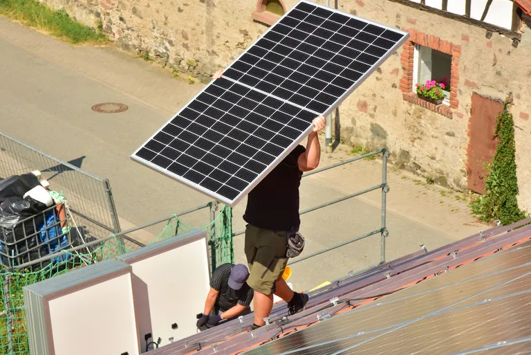 Dachdecker beim Aufbau einer Photovoltaikanlage auf einem neu gedeckten Ziegeldach: Das Schienensystem ist montiert und die Paneele werden an den Verlegeort getragen.