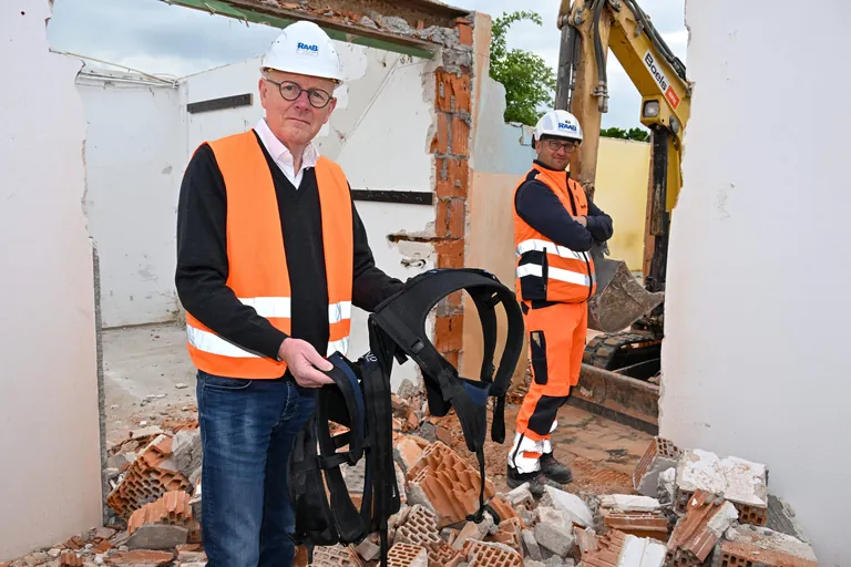 Wolfgang Schubert-Raab steht auf der Baustelle mit einem Exoskelett in den Händen, sein Polier Florian Esselberger steht im Hintergrund
