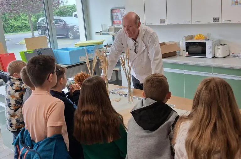 Ein B&auml;cker im wei&szlig;en B&auml;ckerhemd erkl&auml;rt einer Gruppe von Kindern Getreide.