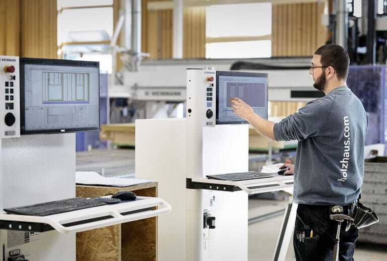 Ein Mann steht vor einem Bildschirm in einer Fertigungshalle f&uuml;r Holzh&auml;user.